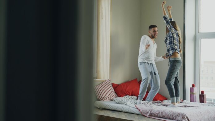 Photo by Vitaly Gariev Couple dancing joyfully on a bed near window.